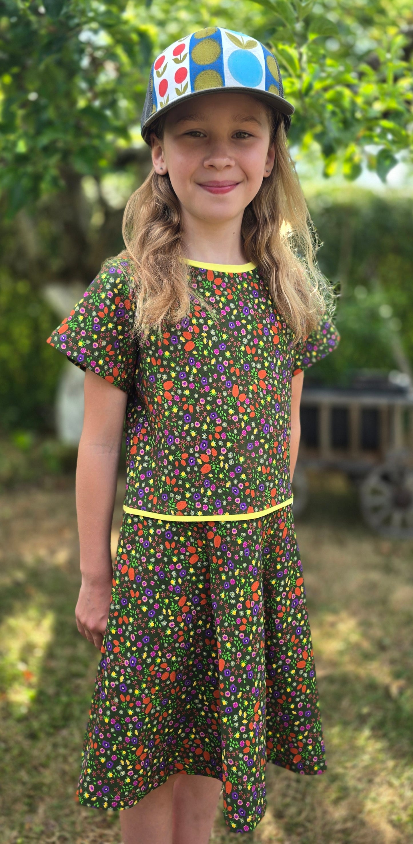Ausgestelltes Kinderkleid mit Millefleur Muster in Oliv grundig, abgesetzt mit gelbem Kantenband kurzarm, verspielt.