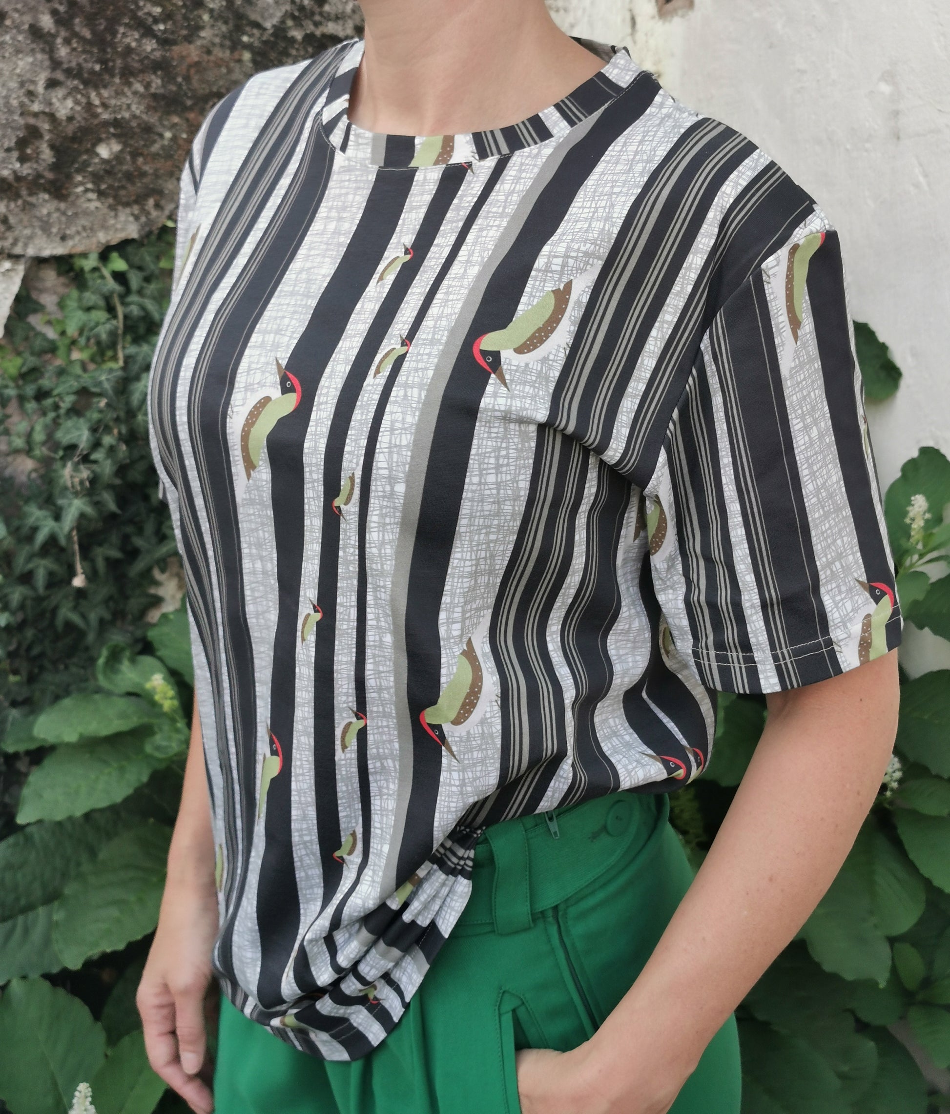 T-Shirt aus Biobaumwolle mit Streifenmuster in unregelmäßigen Abständen- die Streifen sind schwarz auf weiß und ein Grünspecht pickt Löcher in die Streifen, witzig, verspielt.