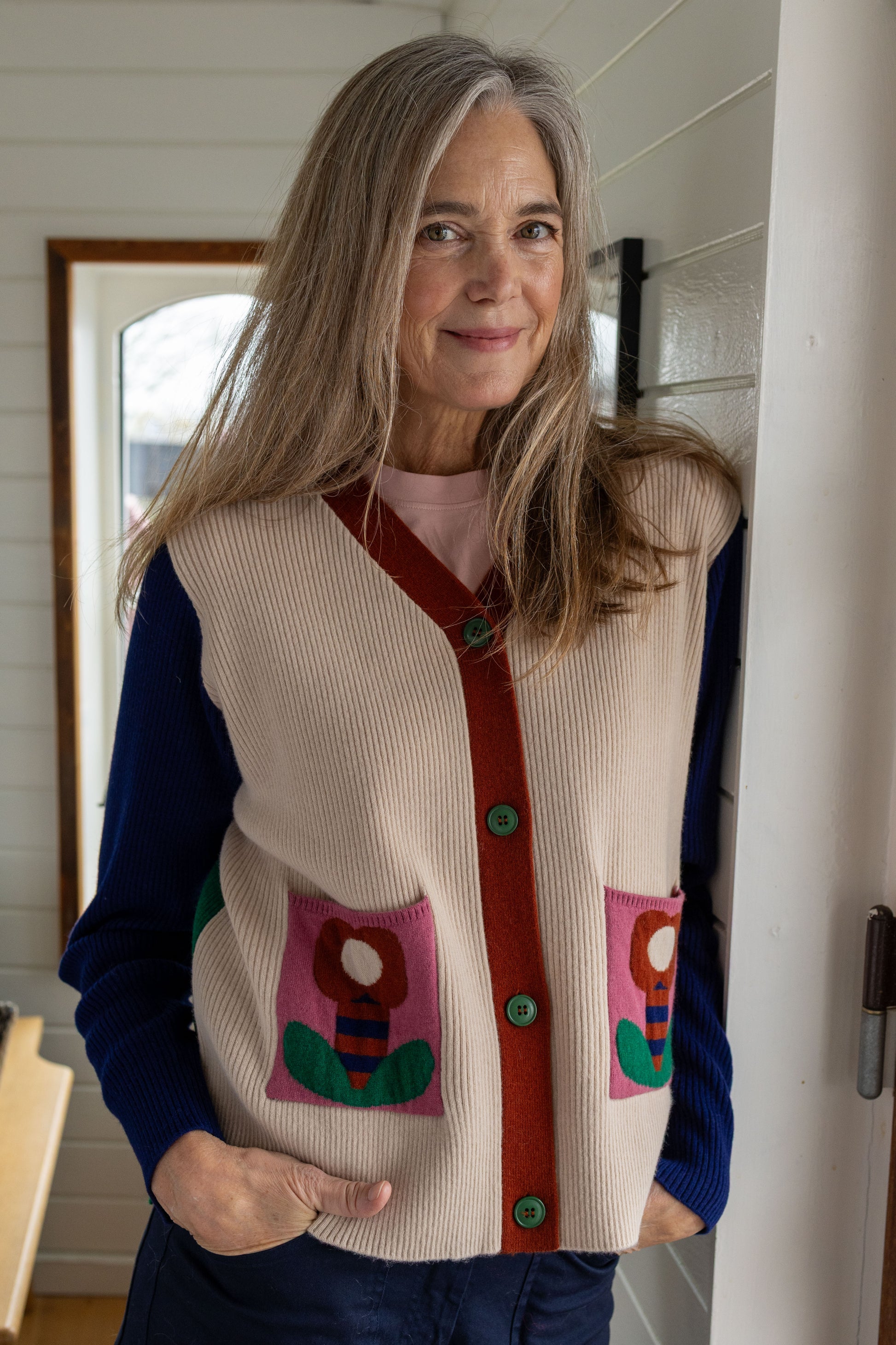 Kassischer, geknöpfter Cardigan mit V-Ausschnitt im Colourblock mit blau Ärmeln, grün Rücken und cremeweiss Vorderteil, die kleinen Aufsatztaschen frontal sorgen dann in Pink mit Prilblume fürs Abrunden des herrlichen Retro Charme, Merinowolle sorgt für Wertigkeit dazu.