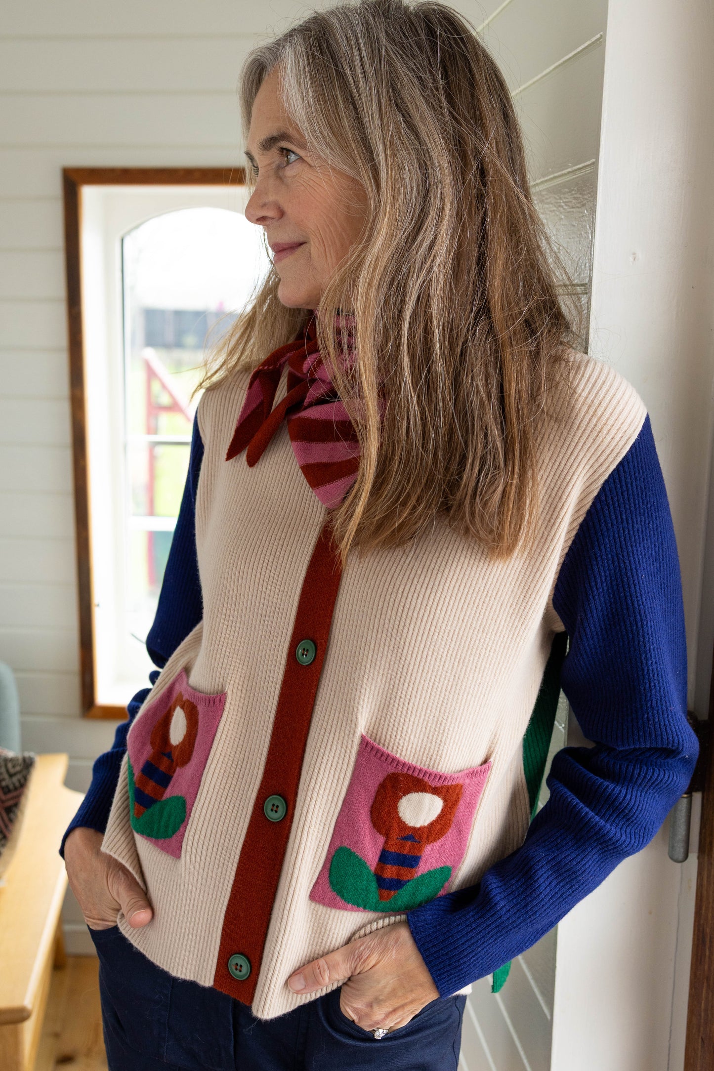 Kassischer, geknöpfter Cardigan mit V-Ausschnitt im Colourblock mit blau Ärmeln, grün Rücken und cremeweiss Vorderteil, die kleinen Aufsatztaschen frontal sorgen dann in Pink mit Prilblume fürs Abrunden des herrlichen Retro Charme, Merinowolle sorgt für Wertigkeit dazu.