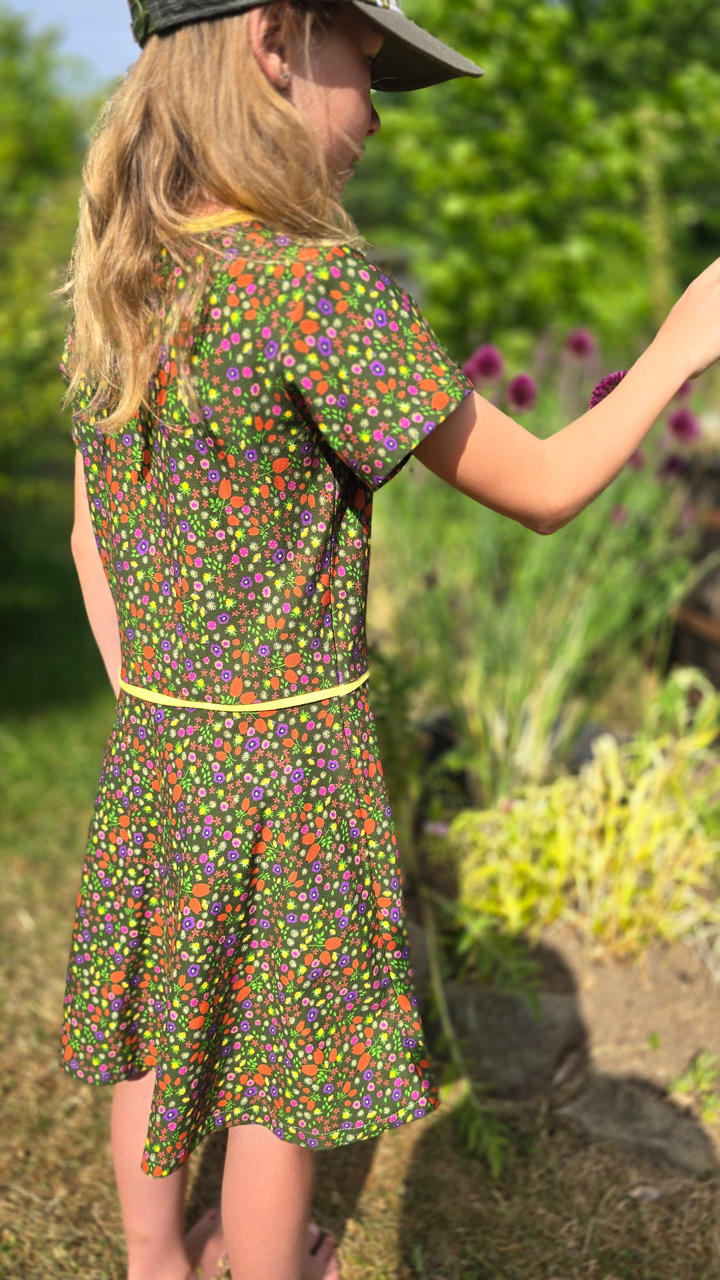Ausgestelltes Kinderkleid mit Millefleur Muster in Oliv grundig, abgesetzt mit gelbem Kantenband kurzarm, verspielt.